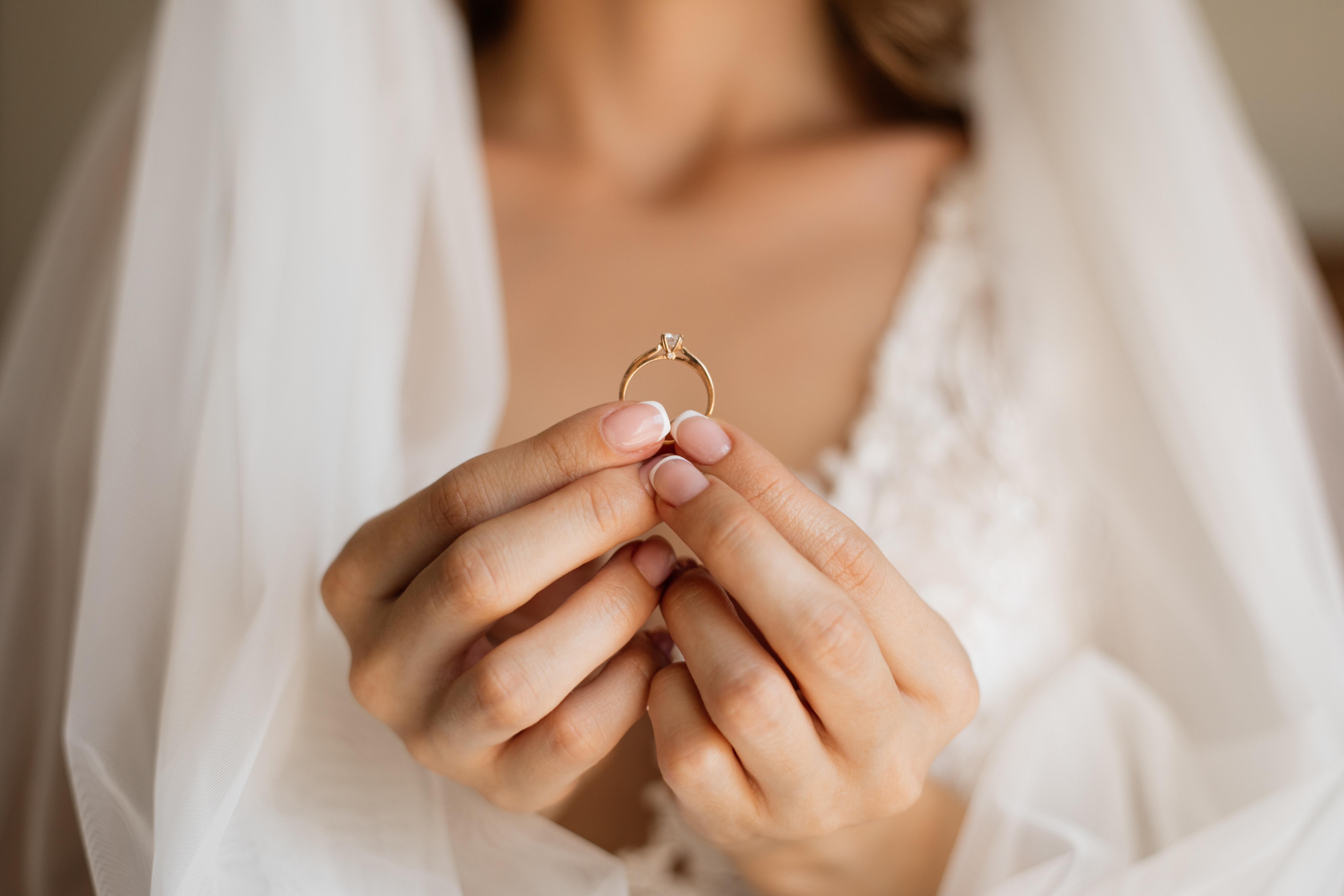 Front view of bride is holding precious engagement ring in hands in front of chest and wedding veil